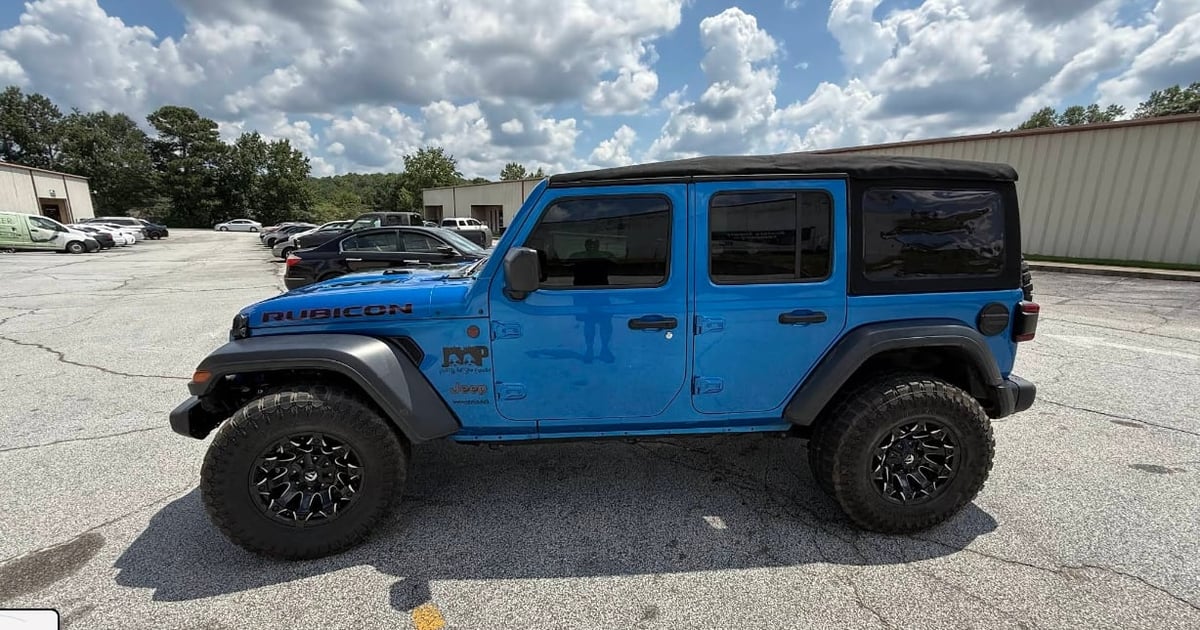 Bright blue Jeep Wrangler four-door SUV with black wheels and bumpers parked in an outdoor lot under cloudy skies
